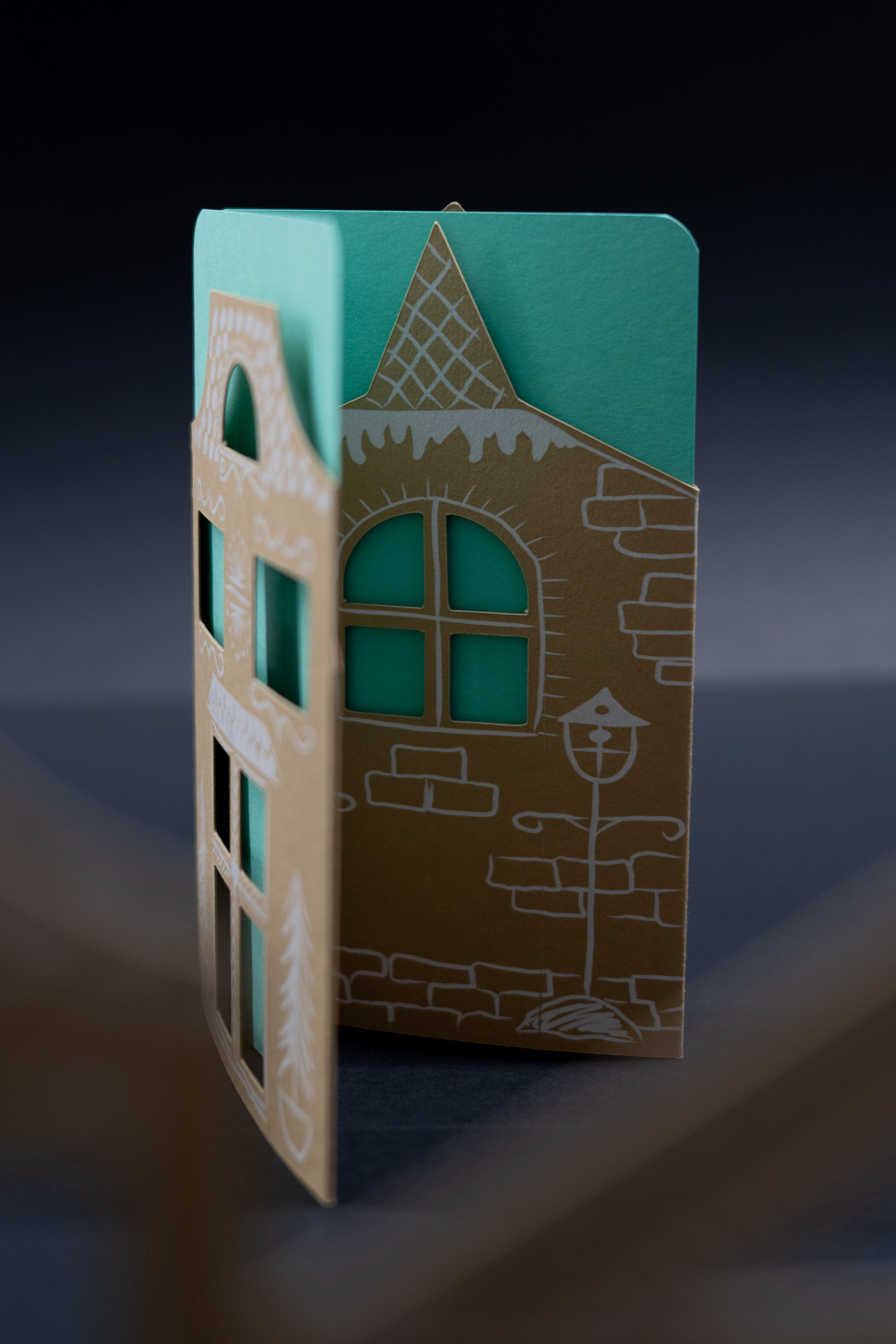 Handgefertigte Papierkarte mit einem Ausschnittdesign, das einer Gebäudefassade mit Fenstern ähnelt, und weißen dekorativen Skizzen auf braunem und blaugrünem Papier.